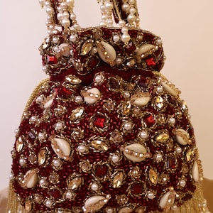 May include: A burgundy beaded handbag with a drawstring closure. The bag is adorned with gold and red beads, faux pearls, and seashells. The handle is made of gold beads and faux pearls.