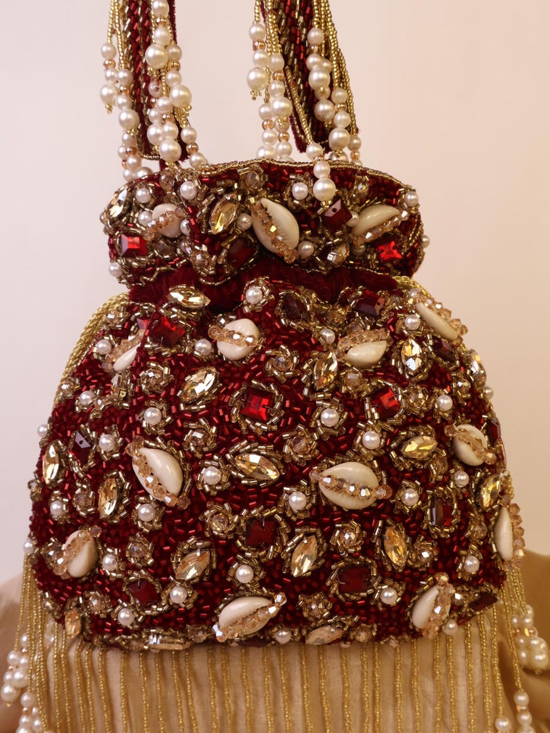 May include: A burgundy beaded handbag with a drawstring closure. The bag is adorned with gold and red beads, faux pearls, and seashells. The handle is made of gold beads and faux pearls.