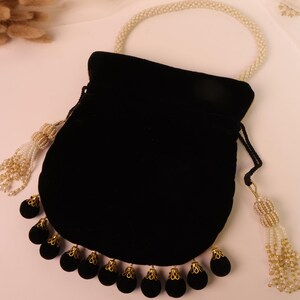 May include: A black velvet drawstring bag with a pearl handle and tassel accents. The bag features a row of black velvet ornaments with gold-coloured filigree caps along the bottom edge. The handle and tassels are adorned with small pearls.