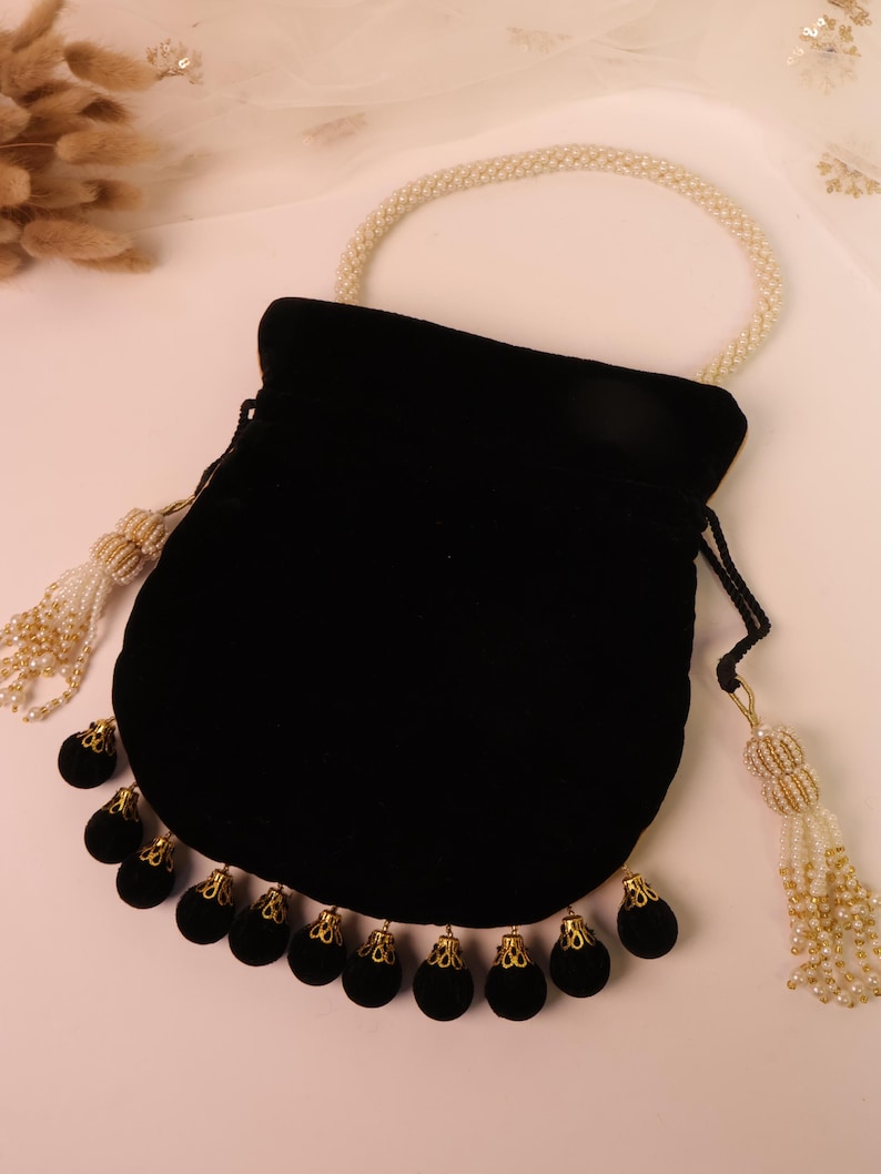 May include: A black velvet drawstring bag with a pearl handle and tassel accents. The bag features a row of black velvet ornaments with gold-coloured filigree caps along the bottom edge. The handle and tassels are adorned with small pearls.