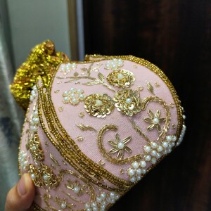 May include: A pink, ornate clutch with gold embroidery and pearl accents. The clutch features floral and scroll designs, with a gold beaded trim and a gold tassel. The bag is likely for special occasions.