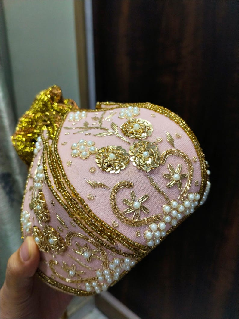 May include: A pink, ornate clutch with gold embroidery and pearl accents. The clutch features floral and scroll designs, with a gold beaded trim and a gold tassel. The bag is likely for special occasions.