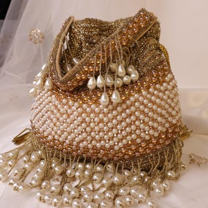 May include: A gold beaded handbag with a drawstring closure. The bag is adorned with white pearls and gold bead fringe. The top and handle are embellished with gold beads and small pearls. The bag is on a white surface.
