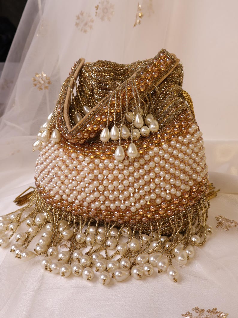 May include: A gold beaded handbag with a drawstring closure. The bag is adorned with white pearls and gold bead fringe. The top and handle are embellished with gold beads and small pearls. The bag is on a white surface.