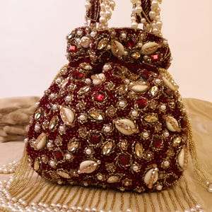 May include: A burgundy beaded handbag with a rounded shape, adorned with red gems, gold accents, and white pearls. The bag features a beaded handle and a fringe of pearls. Shells are also incorporated into the design.