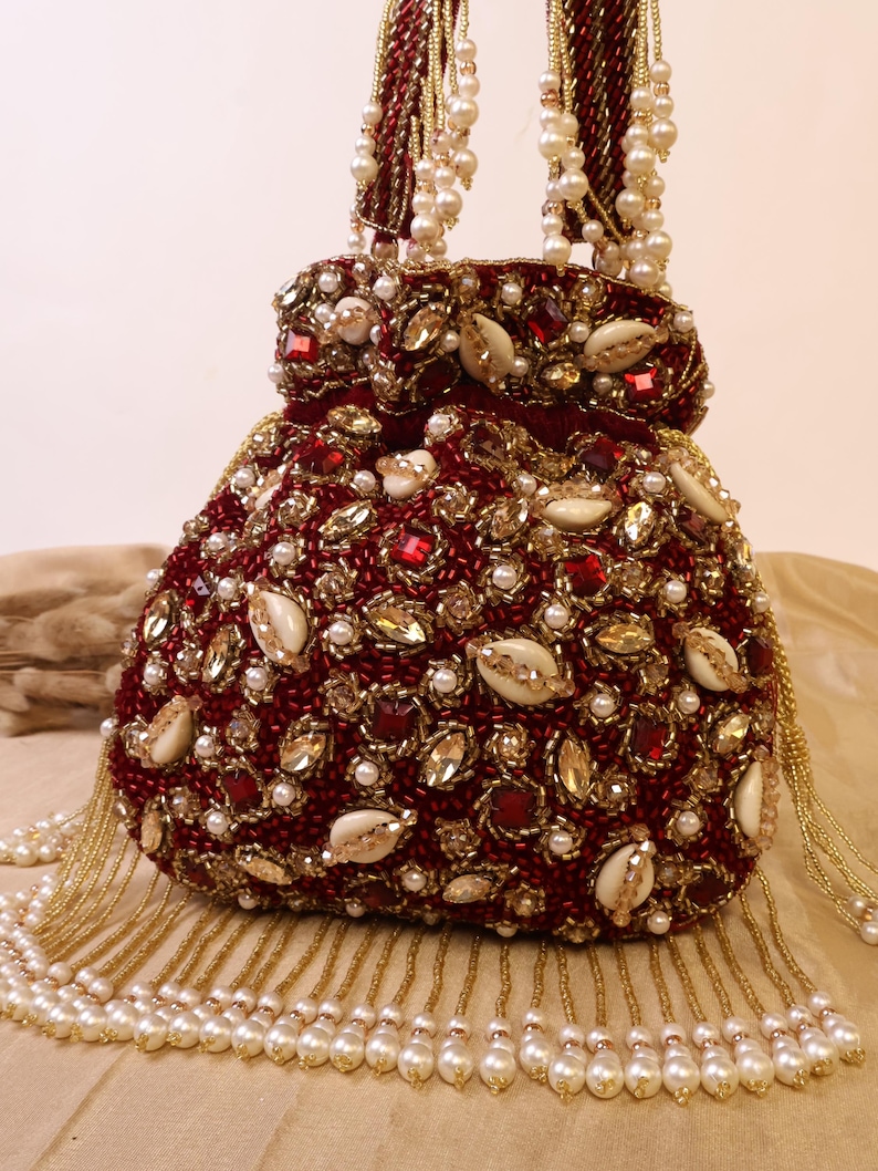 May include: A burgundy beaded handbag with a rounded shape, adorned with red gems, gold accents, and white pearls. The bag features a beaded handle and a fringe of pearls. Shells are also incorporated into the design.