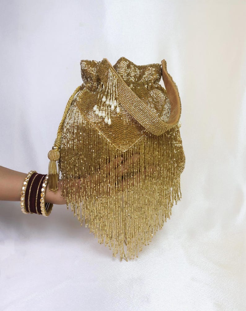 May include: A gold beaded drawstring bag with a matching strap and tassel. The bag is embellished with hanging beads and a decorative flap. A person's arm is visible, adorned with maroon and gold bangles.