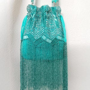 May include: A turquoise drawstring handbag featuring dense beadwork and sequins in chevron patterns. The bag includes a matching turquoise velvet strap and a long, dangling fringe made of thin, delicate turquoise beads covering the lower section.