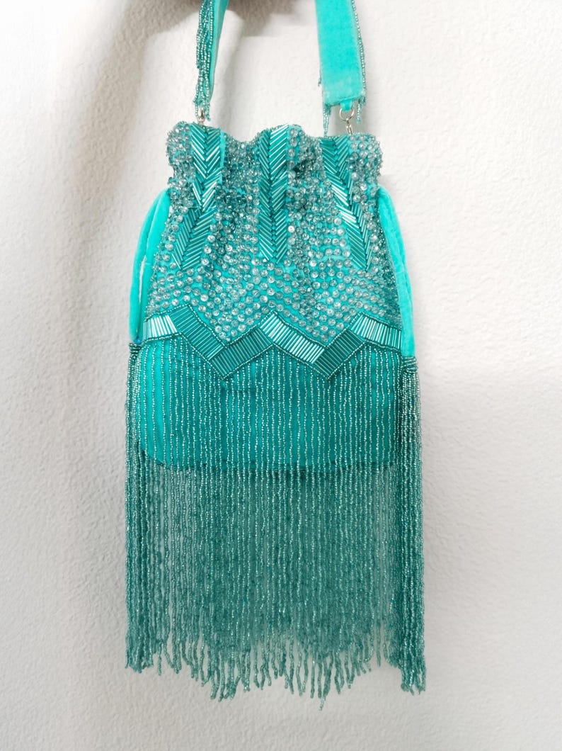 May include: A turquoise drawstring handbag featuring dense beadwork and sequins in chevron patterns. The bag includes a matching turquoise velvet strap and a long, dangling fringe made of thin, delicate turquoise beads covering the lower section.