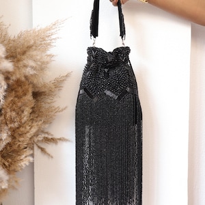 May include: A black beaded handbag featuring a drawstring closure, intricate geometric beadwork on the upper section, and a long, dense fringe that hangs from the base. The bag includes a matching beaded handle attached with silver-toned hardware.