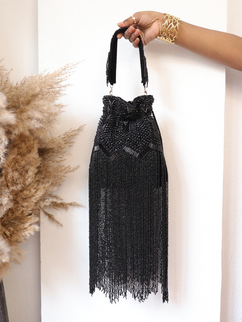 May include: A black beaded handbag featuring a drawstring closure, intricate geometric beadwork on the upper section, and a long, dense fringe that hangs from the base. The bag includes a matching beaded handle attached with silver-toned hardware.