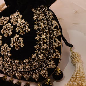 May include: Black velvet drawstring bag with gold and pearl bead embellishments. The bag has a decorative tassel and small black pom-poms. The design incorporates floral and geometric patterns, creating an ornate appearance.