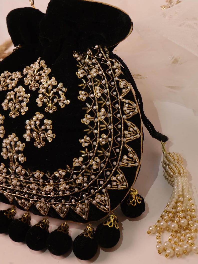 May include: Black velvet drawstring bag with gold and pearl bead embellishments. The bag has a decorative tassel and small black pom-poms. The design incorporates floral and geometric patterns, creating an ornate appearance.