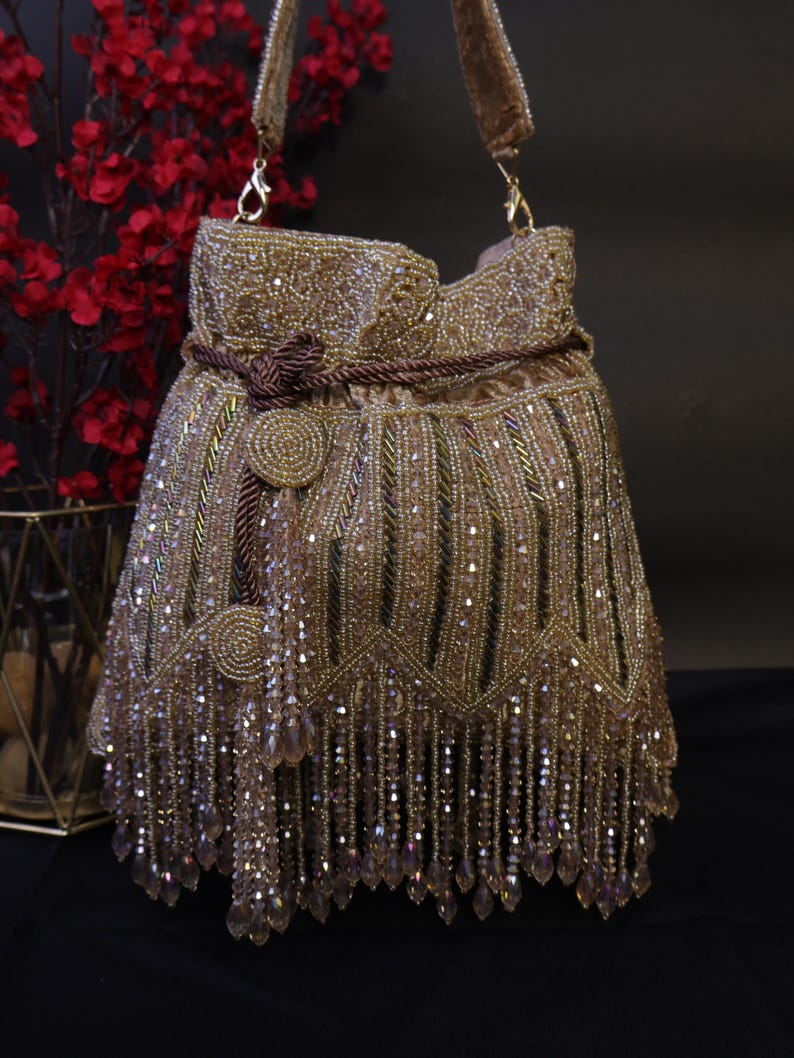 May include: A gold beaded handbag with a drawstring closure and a shoulder strap. The bag features intricate beadwork, a decorative knot, and dangling crystal fringe. The bag is a stylish accessory for special occasions.