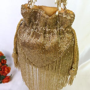 May include: A gold beaded drawstring bag with a tassel fringe. The bag features a gold chain strap with pearl accents and a gold lining. The bag is embellished with gold beads and has a decorative tassel on the side.