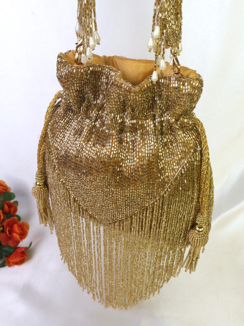 May include: A gold beaded drawstring bag with a tassel fringe. The bag features a gold chain strap with pearl accents and a gold lining. The bag is embellished with gold beads and has a decorative tassel on the side.