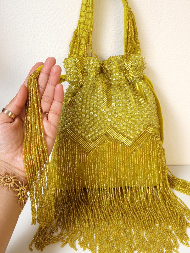 May include: A chartreuse drawstring handbag featuring intricate beadwork and sequins in a chevron pattern. The bag includes a beaded handle and long, dangling beaded fringe along the bottom edge. The drawstring closure is accented with beaded tassels.