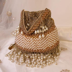 May include: A beaded drawstring bag in shades of gold and cream. The bag is covered in small, round beads, with a pattern of cream-colored beads and gold-colored beads. The bottom edge is trimmed with dangling pearl-shaped beads and gold fringe.