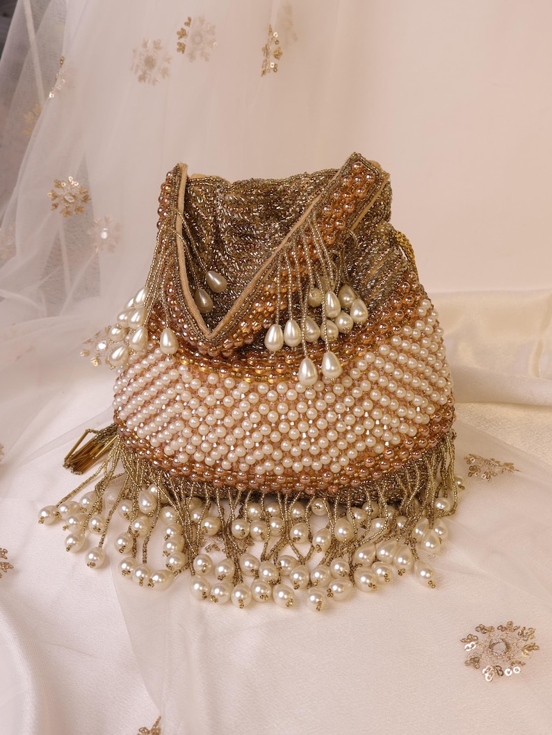 May include: A beaded drawstring bag in shades of gold and cream. The bag is covered in small, round beads, with a pattern of cream-colored beads and gold-colored beads. The bottom edge is trimmed with dangling pearl-shaped beads and gold fringe.