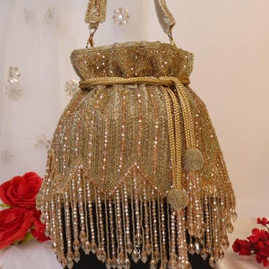 May include: A gold beaded handbag with a drawstring closure and a matching handle. The bag features intricate beadwork and dangling crystal-like embellishments, creating a luxurious and ornate appearance. The bag is approximately 25 cm tall.