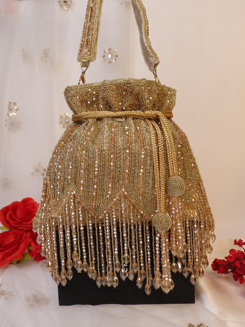May include: A gold beaded handbag with a drawstring closure and a matching handle. The bag features intricate beadwork and dangling crystal-like embellishments, creating a luxurious and ornate appearance. The bag is approximately 25 cm tall.