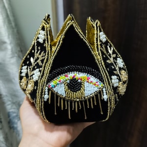May include: Black velvet clutch with a gold beaded trim and a large beaded eye design. The eye features colorful accents and gold teardrop details. The clutch has a unique, petal-like shape and is embellished with white floral accents.