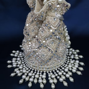 Peut inclure: Un sac à cordon de serrage ivoire, orné de perles, de paillettes et de strass carrés. Le sac présente une frange de perles à la base et une poignée perlée. Le sac est posé sur un fond bleu foncé. Le design est de style vintage.