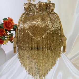 May include: A gold beaded drawstring bag with a triangular fringe. The bag has a rounded top with beaded handles and tassels. The bag is made of small, shiny gold beads.
