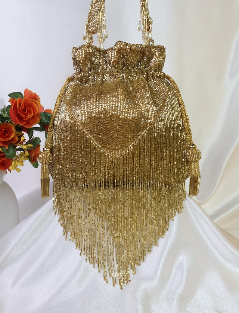 May include: A gold beaded drawstring bag with a triangular fringe. The bag has a rounded top with beaded handles and tassels. The bag is made of small, shiny gold beads.