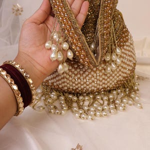 May include: A beaded clutch with a gold handle and pearl accents. The bag is covered in small, off-white beads and features a fringe of dangling pearls. A maroon and gold bracelet is visible on the left.