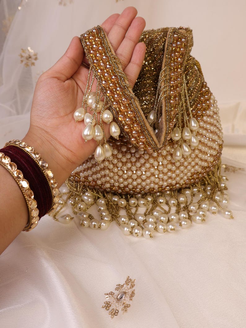 May include: A beaded clutch with a gold handle and pearl accents. The bag is covered in small, off-white beads and features a fringe of dangling pearls. A maroon and gold bracelet is visible on the left.