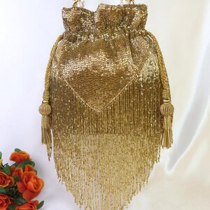 May include: A gold beaded drawstring bag with a diamond-shaped design on the front. The bag features long beaded fringe and gold rope handles with tassels. The bag is a glamorous accessory.