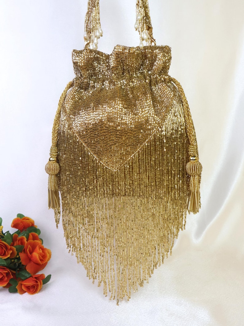 May include: A gold beaded drawstring bag with a diamond-shaped design on the front. The bag features long beaded fringe and gold rope handles with tassels. The bag is a glamorous accessory.