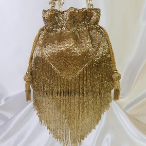 May include: A gold beaded drawstring bag with a triangular fringe. The bag features a gathered top, beaded handles, and decorative tassels. The bag is made of small, shiny gold beads.
