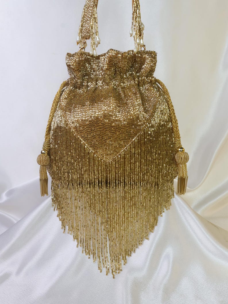 May include: A gold beaded drawstring bag with a triangular fringe. The bag features a gathered top, beaded handles, and decorative tassels. The bag is made of small, shiny gold beads.