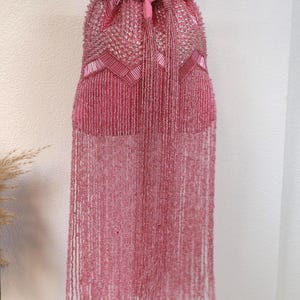 May include: A pink drawstring handbag featuring dense beadwork in a chevron pattern across the upper section. The bag includes a matching pink fabric tie closure and a long, flowing fringe made of thin, delicate pink beaded strands.