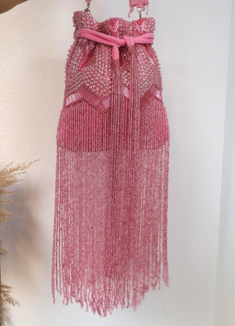 May include: A pink drawstring handbag featuring dense beadwork in a chevron pattern across the upper section. The bag includes a matching pink fabric tie closure and a long, flowing fringe made of thin, delicate pink beaded strands.