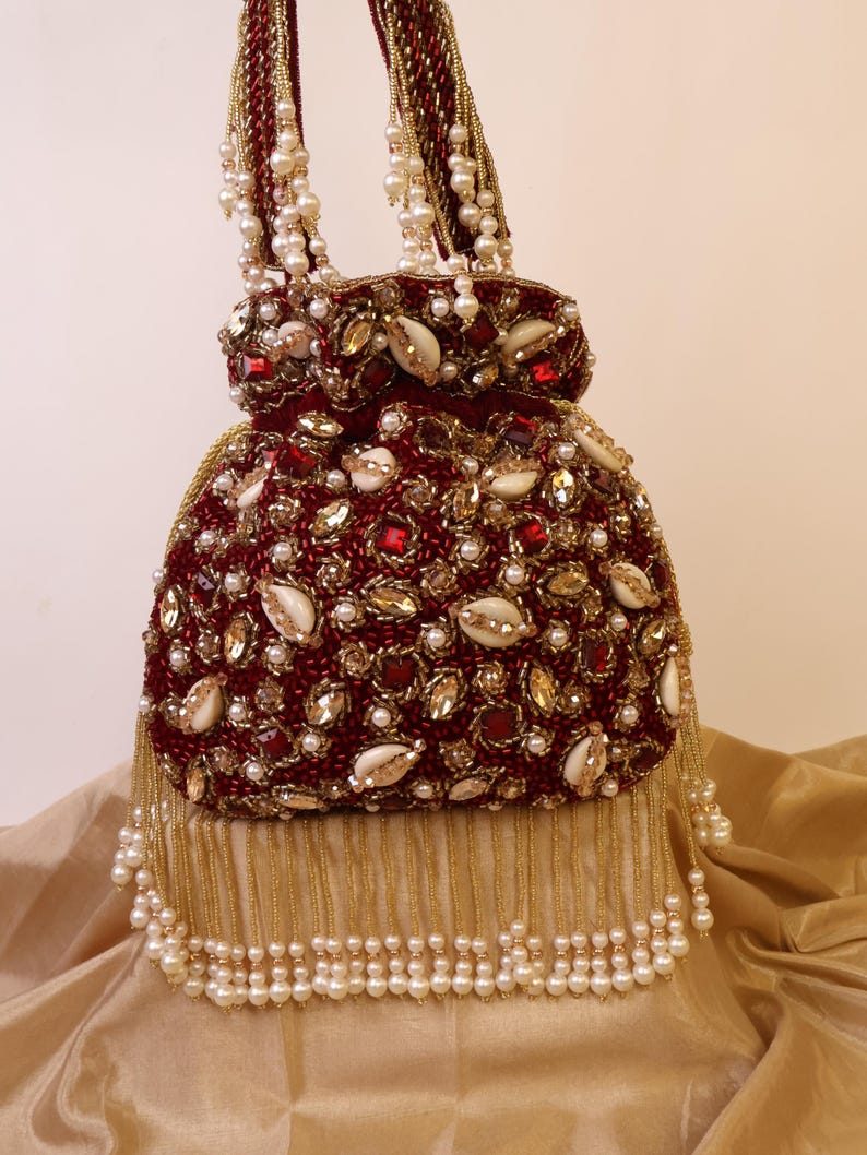 May include: A burgundy beaded handbag with gold and pearl accents. The bag features a rounded shape, embellished with red gems, gold sequins, and white shell-shaped details. Pearl strands form the handle and a decorative fringe.
