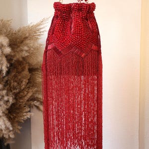 May include: A vibrant red drawstring handbag featuring intricate beadwork and long, flowing beaded fringe. The upper section displays a geometric pattern of glass beads and tube beads, while the lower portion consists of dense, hanging red fringe.