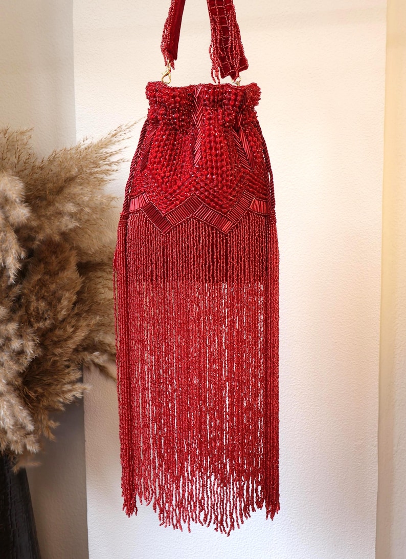 May include: A vibrant red drawstring handbag featuring intricate beadwork and long, flowing beaded fringe. The upper section displays a geometric pattern of glass beads and tube beads, while the lower portion consists of dense, hanging red fringe.