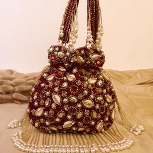 May include: A burgundy beaded handbag with gold and pearl accents. The bag features a rounded shape, embellished with red and gold stones, white shells, and small pearls. The handles and tassels are adorned with gold beads and white pearls.