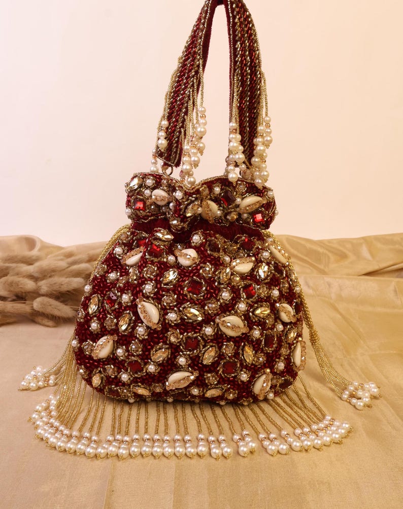 May include: A burgundy beaded handbag with gold and pearl accents. The bag features a rounded shape, embellished with red and gold stones, white shells, and small pearls. The handles and tassels are adorned with gold beads and white pearls.