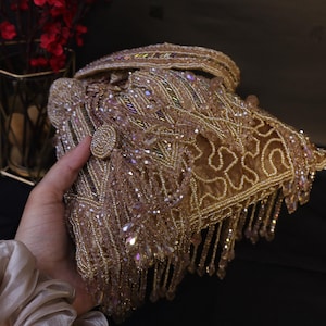 May include: A gold beaded clutch bag with detailed embellishments and dangling fringe. The bag has a button closure and is decorated with iridescent crystals. A glamorous accessory for special occasions.