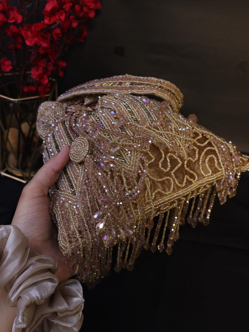 May include: A gold beaded clutch bag with detailed embellishments and dangling fringe. The bag has a button closure and is decorated with iridescent crystals. A glamorous accessory for special occasions.