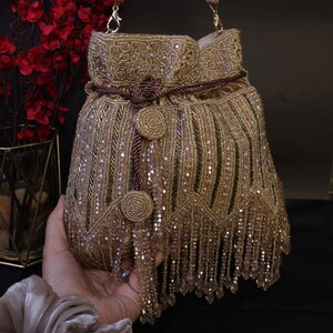May include: A gold beaded handbag with a drawstring closure and a shoulder strap. The bag features intricate beadwork, a tassel fringe, and decorative circular embellishments. The bag is held by a hand.