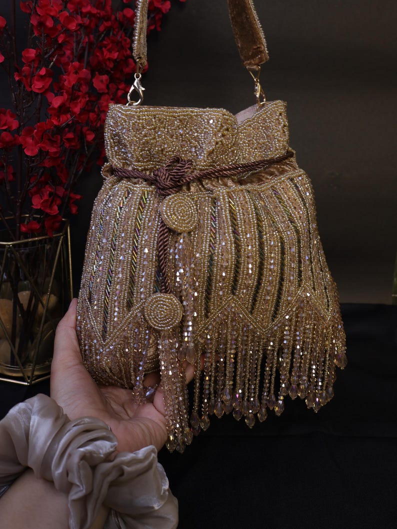 May include: A gold beaded handbag with a drawstring closure and a shoulder strap. The bag features intricate beadwork, a tassel fringe, and decorative circular embellishments. The bag is held by a hand.