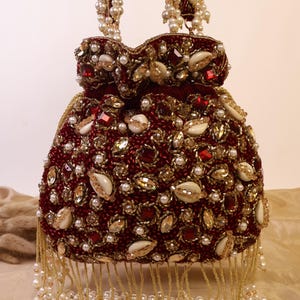 May include: A burgundy beaded handbag with pearl and gold accents. The bag features a drawstring closure, embellished with pearls, gemstones, and seashells. The handles are adorned with pearl strands, and the bottom has a fringe of pearls.