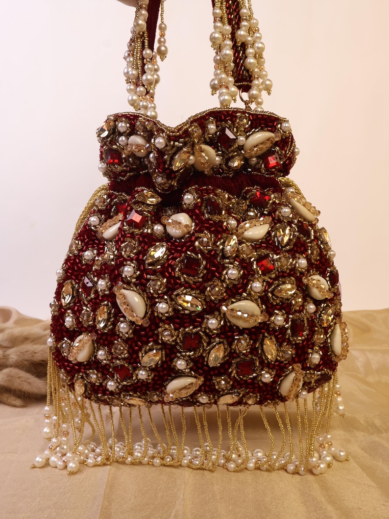 May include: A burgundy beaded handbag with pearl and gold accents. The bag features a drawstring closure, embellished with pearls, gemstones, and seashells. The handles are adorned with pearl strands, and the bottom has a fringe of pearls.