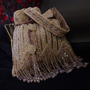 Könnte beinhalten: Eine goldfarbene, mit Perlen besetzte Handtasche mit Schulterriemen und Fransen. Die Tasche hat aufwendige Perlenarbeiten, einen Kordelzugverschluss und dekorative kreisförmige Verzierungen. Die Tasche ist hellgoldfarben mit schillernden Perlen.