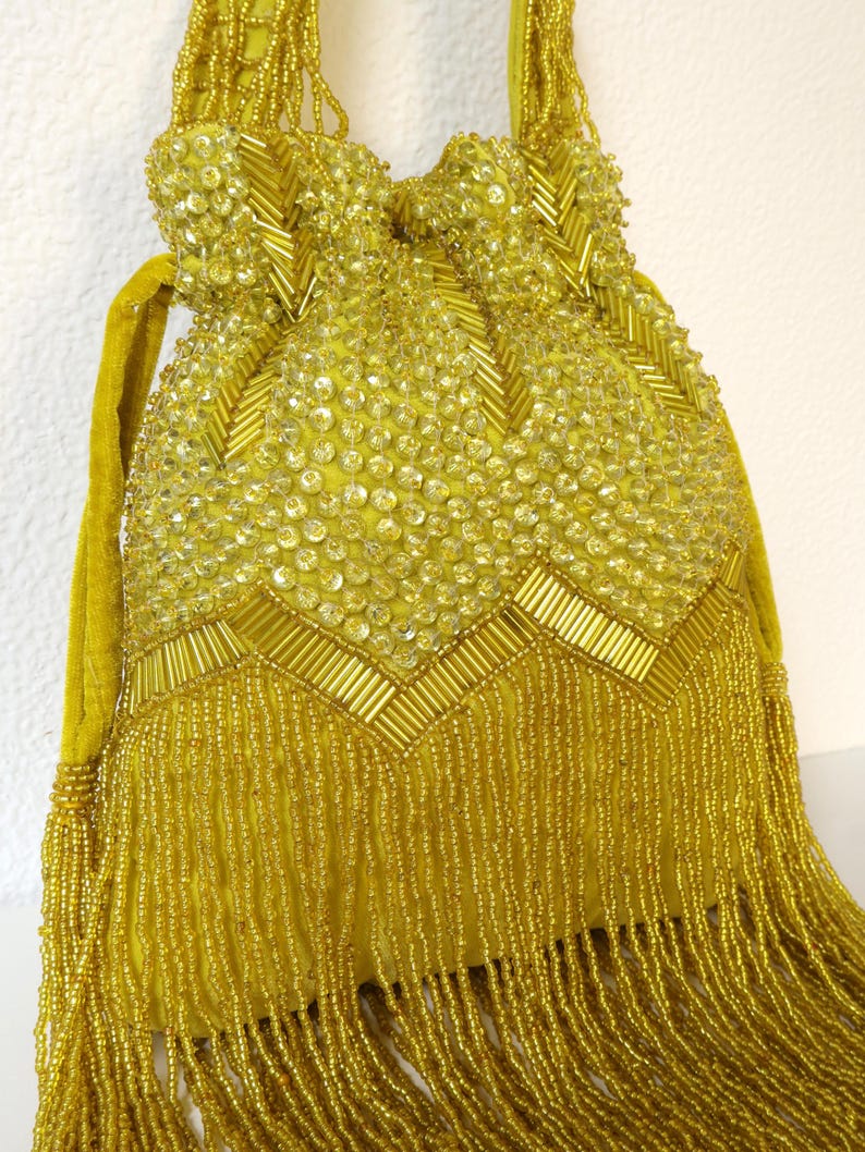 May include: A mustard yellow drawstring pouch bag featuring extensive beadwork and sequin embellishments. The exterior is decorated with a chevron pattern of cylindrical beads and circular sequins, finished with a long fringe of dangling yellow beads.